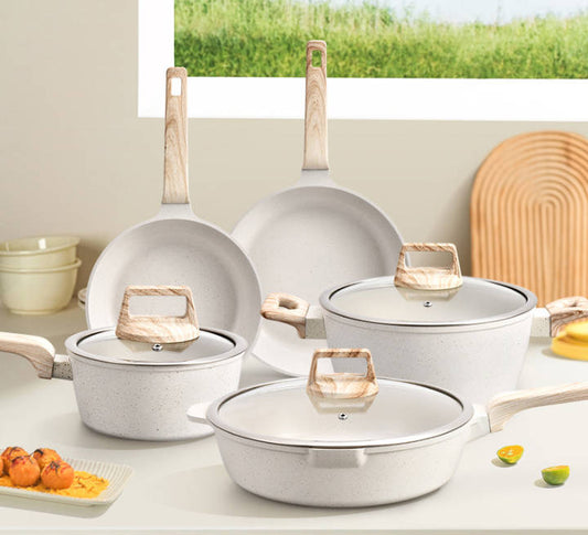 A cookware set with light speckled white ceramic pots and pans, featuring light wood handles and glass lids, arranged on a kitchen counter with a window view in the background.