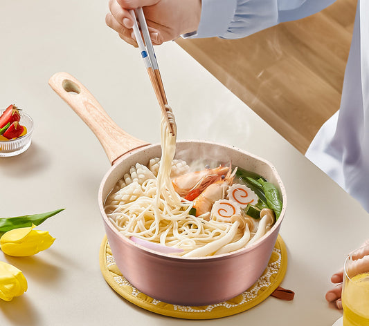 Person using chopsticks to eat noodles from a pink pot on a light surface with flowers and a glass of juice.