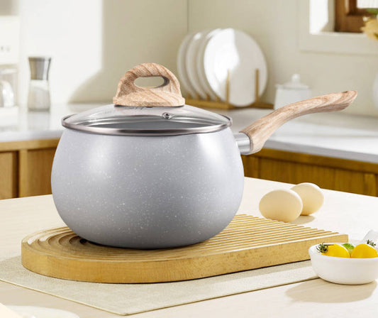 A gray speckled saucepan with a wooden handle and matching lid sits on a wooden trivet in a bright kitchen setting, with eggs and lemons nearby.